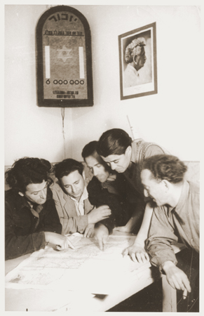 Members of Kibbutz Nili (a Zionist agricultural collective) study a map of Palestine. Above them hangs a wall plaque memorializing the six million Jews killed during the Holocaust. On the other wall is a photograph of the Labor Zionist leader, Berl Katznelson. Pleikershof, Germany, 1945-1948.