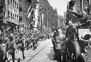 Membros da Juventude Hitlerista marcham na presença do seu líder, Baldur von Schirach (à direita, efetuando a saudação), e de outros oficiais nazistas, incluindo Julius Streicher.  Foto tirada em Nuremberg, Alemanha, 1933.