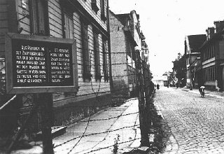 Placa bilíngue, em alemão e letão, avisa que a pessoa que  tentar pular o muro ou entrar em contato com os judeus confinados no gueto de Riga será fuzilada. Foto tirada em Riga, Letônia, 1941-1943.